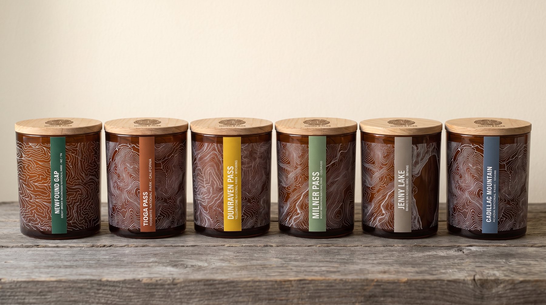 Six Places candles in a row, one from each park: amber jars, white contours, a coloured estate-paper label on each, pine lids