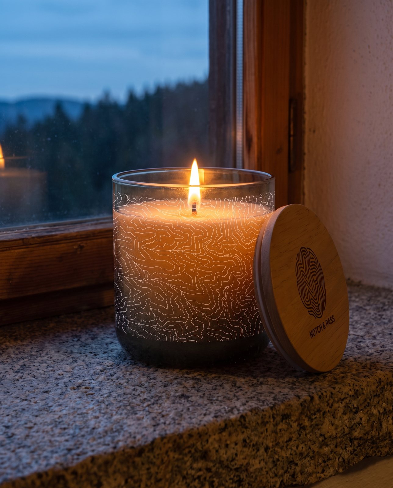 The Woodsmoke & Birch Tar candle lit on a granite sill at dusk, its pine lid leaning beside it, a forested ridge through the window